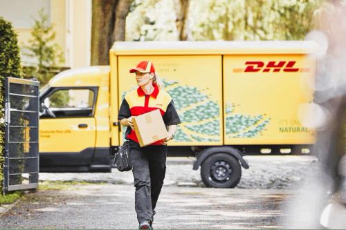  Ein DHL-Zusteller geht mit einem Paket in der Hand über einen Schotterweg. Im Hintergrund parkt ein gelber DHL-Zustellfahrzeug.