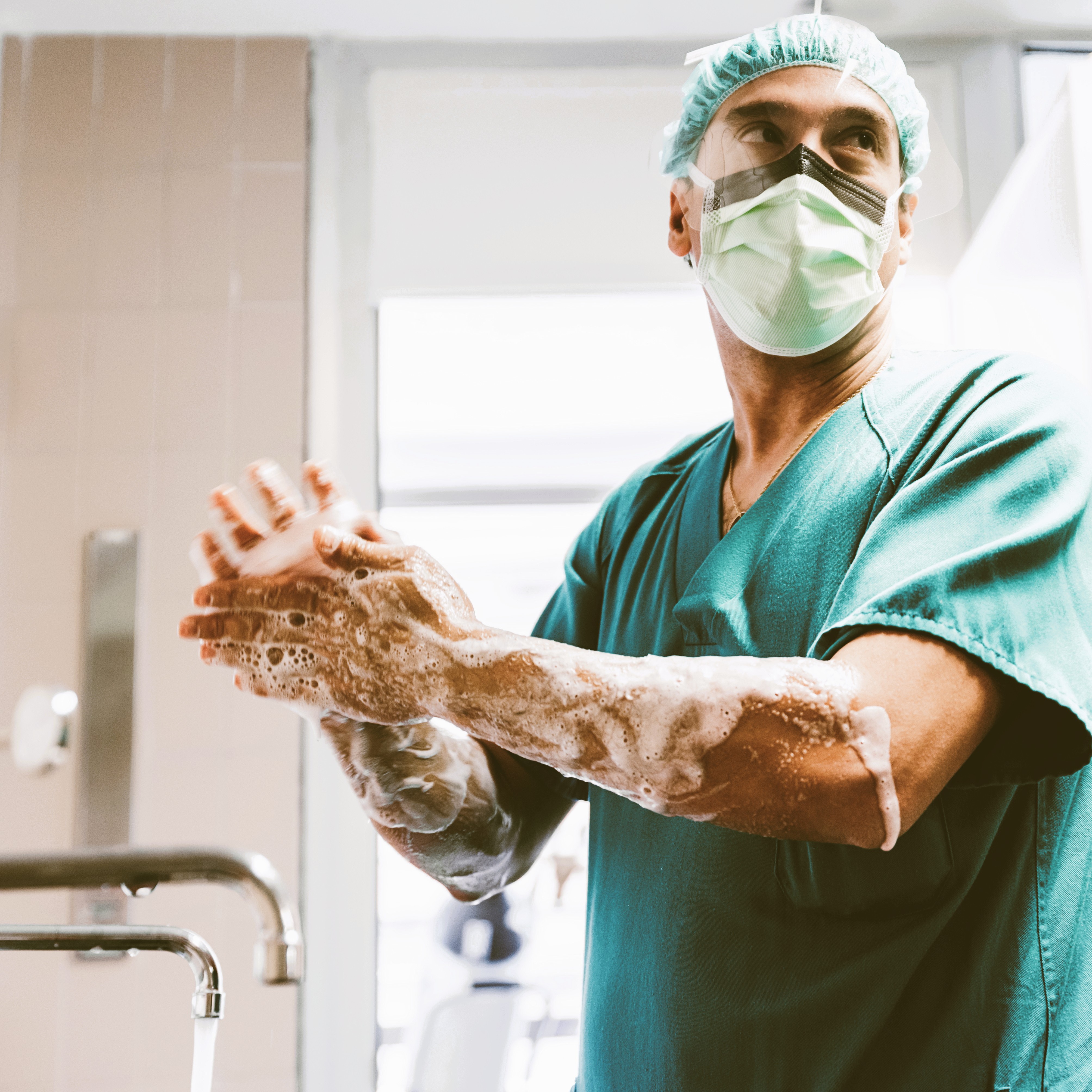 Ein Chirurg in grüner OP-Kleidung und Maske wäscht sich konzentriert die Hände mit Seife an einem Waschbecken, als Vorbereitung auf die Operation