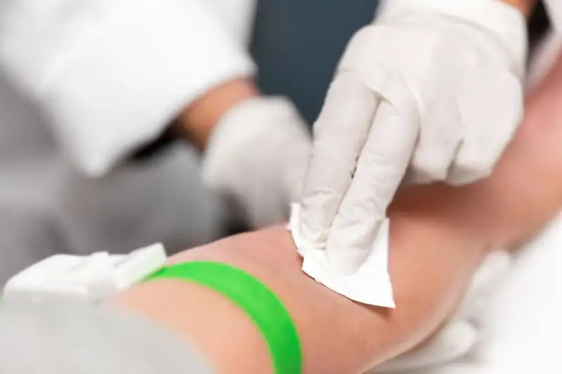 Ein Mitarbeiter des Gesundheitswesens mit weißen Handschuhen reinigt den Arm eines Patienten mit einem Tuch, um eine Blutentnahme vorzubereiten