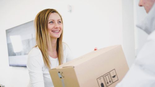  Eine Frau im weißen T-Shirt lächelt, während sie ein Paket von PraxisPanda entgegennimmt. Die Kulisse ist hell, minimalistisch und einladend.